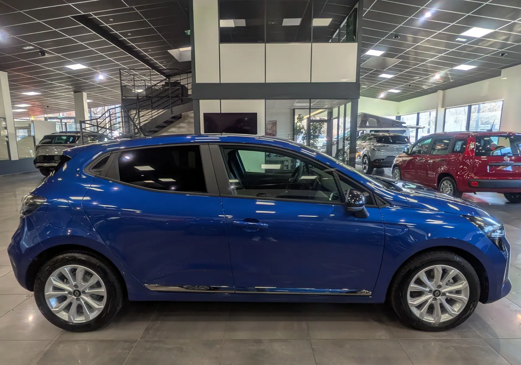 Profil côté gauche d'une Renault Clio V Bleu Iron 2025 en intérieur, avec jantes alliage et vitres teintées.