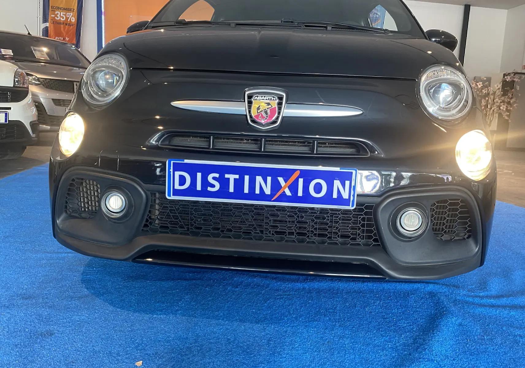 Vue frontale d'une ABARTH 595 noire avec phares allumés et logo Abarth bien visible sur fond intérieur showroom.