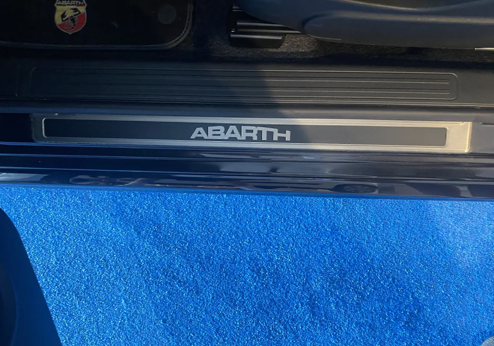 Gros plan sur la plaque de seuil de porte ABARTH noire avec logo sur tapis bleu vif, intérieur côté conducteur visible.