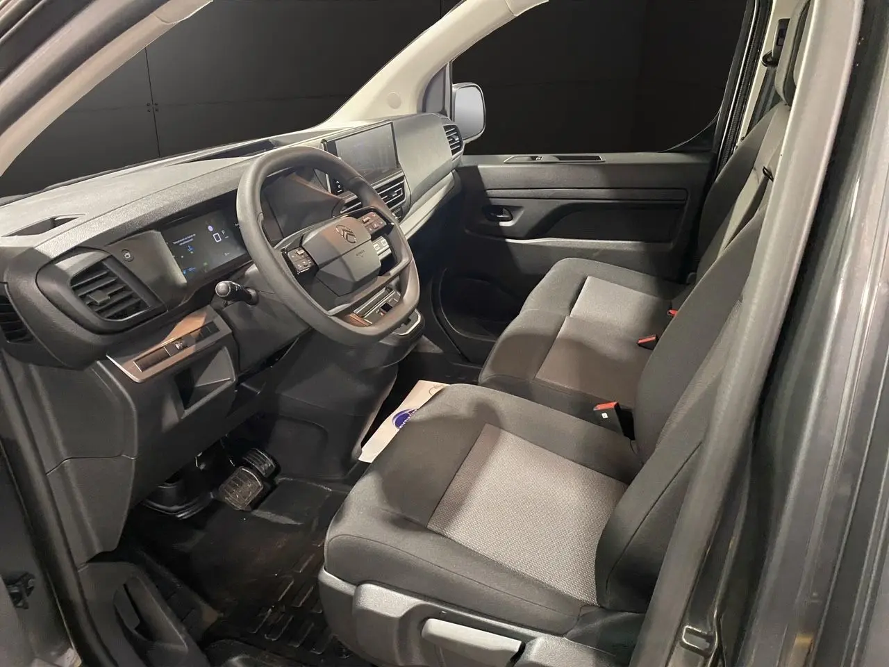 Intérieur du Citroën Jumpy Fourgon 2024 vu côté conducteur, sièges tissu bi-ton gris et tableau de bord moderne.