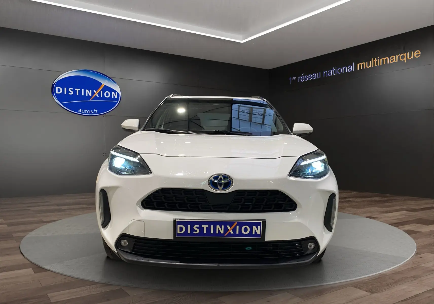 Vue frontale d'une Toyota Yaris Cross blanche avec phares LED allumés dans un showroom moderne.