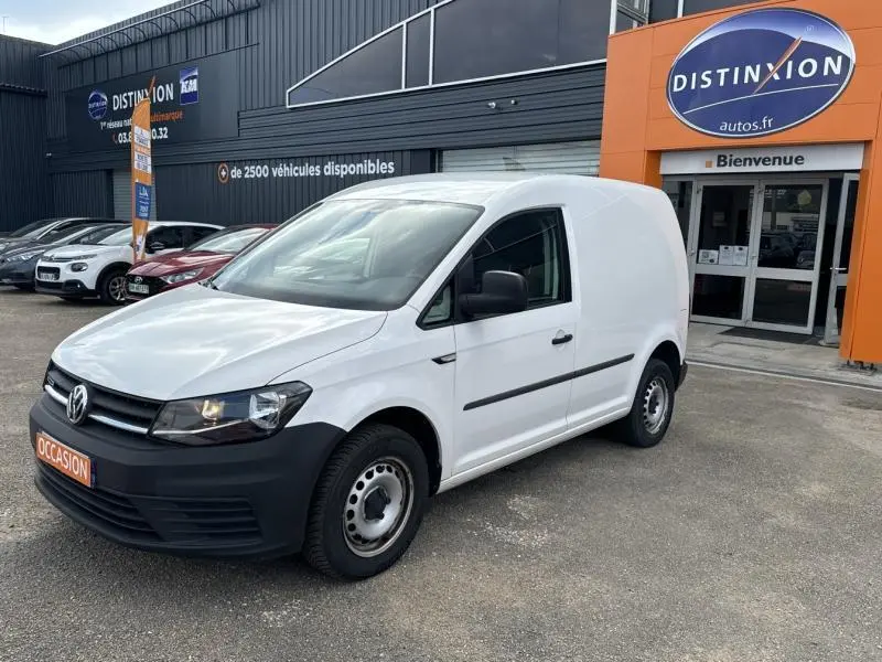 Vue 3/4 avant d'un Volkswagen Caddy Van blanc Candy avec pare-chocs noirs, stationné devant un concessionnaire Distinxion.