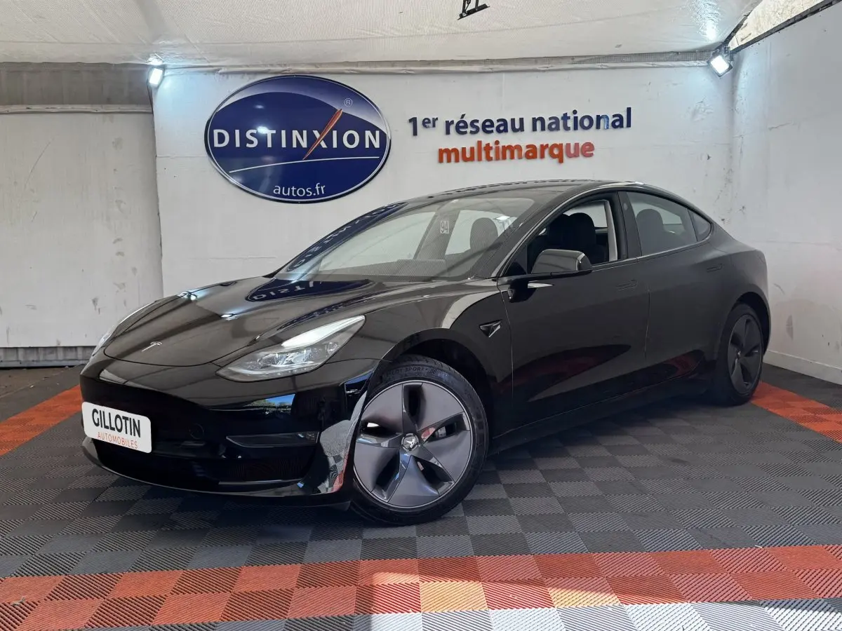 Tesla Model 3 Grande Autonomie noir, vue 3/4 avant droit dans un showroom avec sol à damiers rouge et noir.