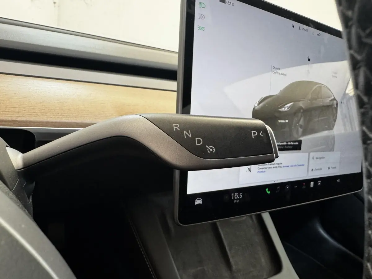 Gros plan sur la commande de boîte automatique noire de la Tesla Model 3 2023, avec écran tactile et tableau de bord bois en arrière-plan.