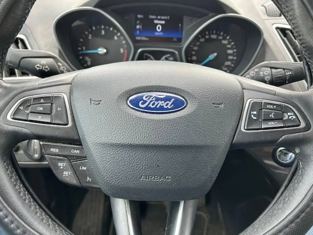 Vue rapprochée du volant cuir noir avec logo Ford et commandes intégrées du tableau de bord d'une Ford C-MAX gris clair.