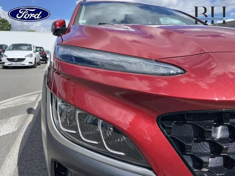 Gros plan sur le phare avant gauche et la calandre d’un Hyundai Kona Pulse Red Métal sous un ciel partiellement nuageux.