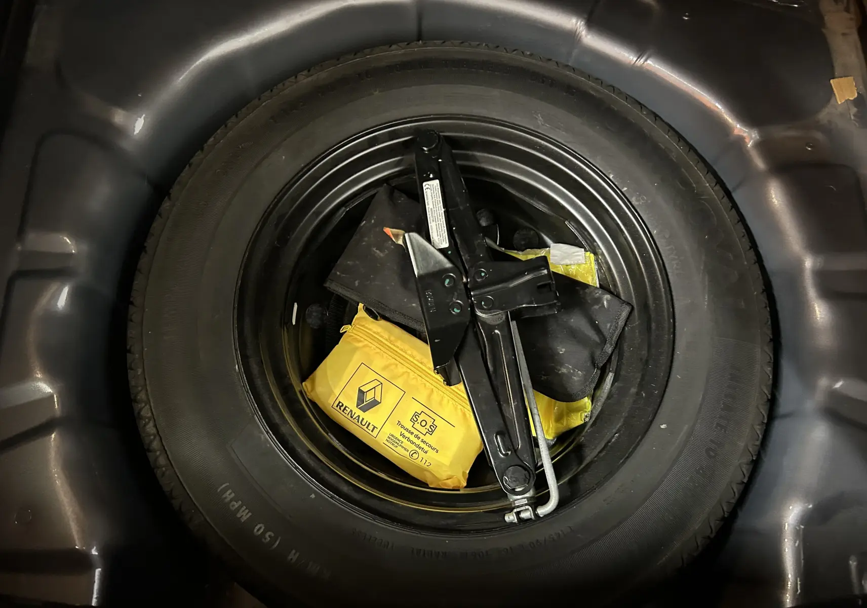 Roue de secours noire avec cric et trousse de secours jaune Renault dans le coffre d'un Renault Captur gris.