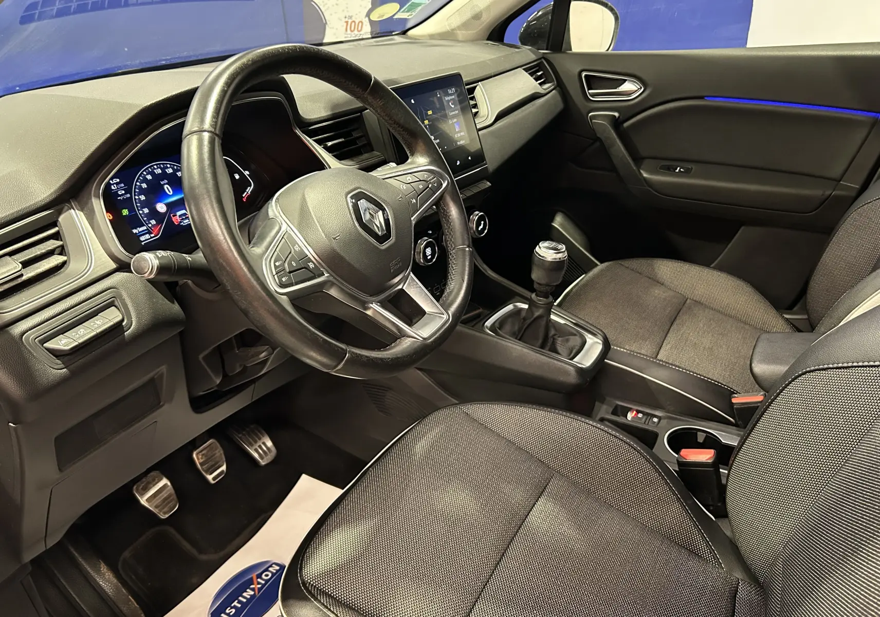 Vue intérieure côté conducteur du Renault Captur gris 2020, volant cuir, boîte manuelle et éclairage d'ambiance bleu sur la porte.