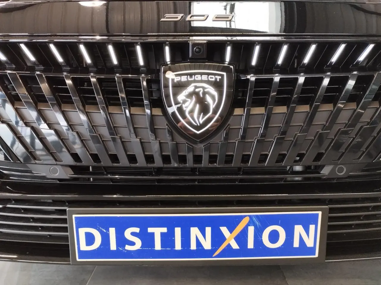 Gros plan sur la calandre noire métallisée d'une Peugeot 308 avec logo lion lumineux et plaque Distinxion.