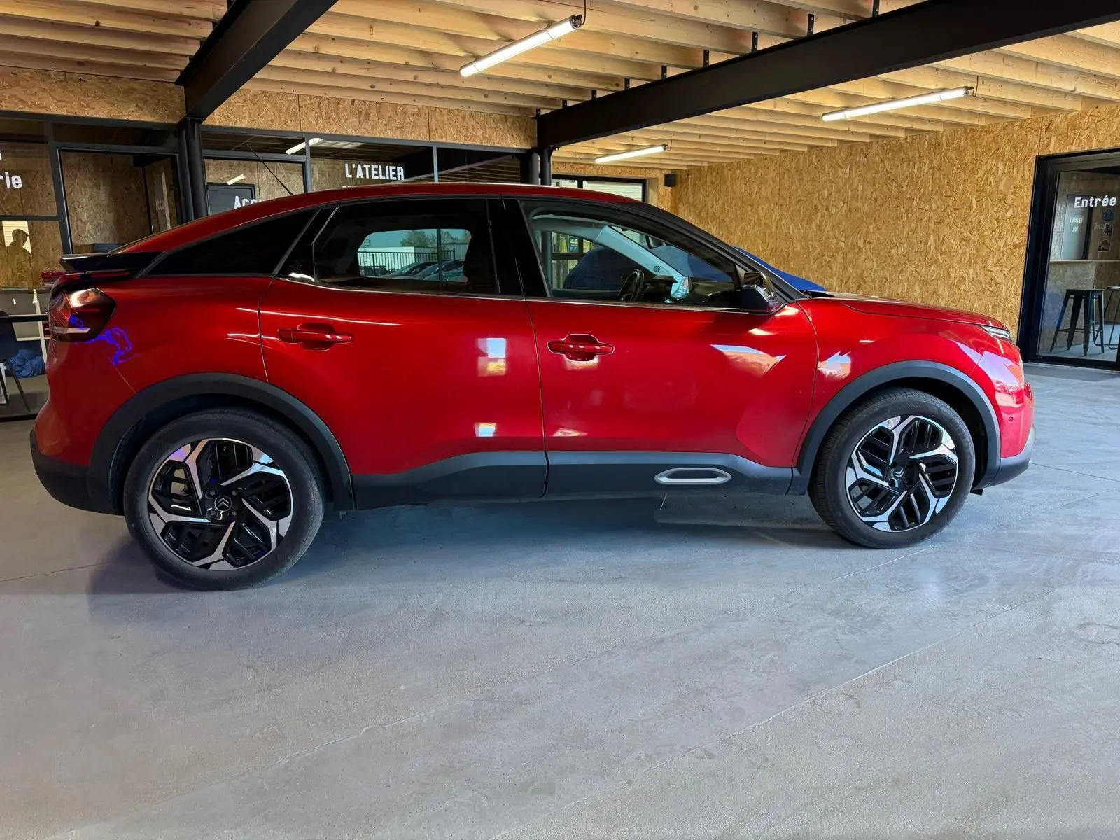 Vue de profil côté gauche d'une Citroën C4 X rouge avec jantes noires et détails noirs sous un abri en bois.