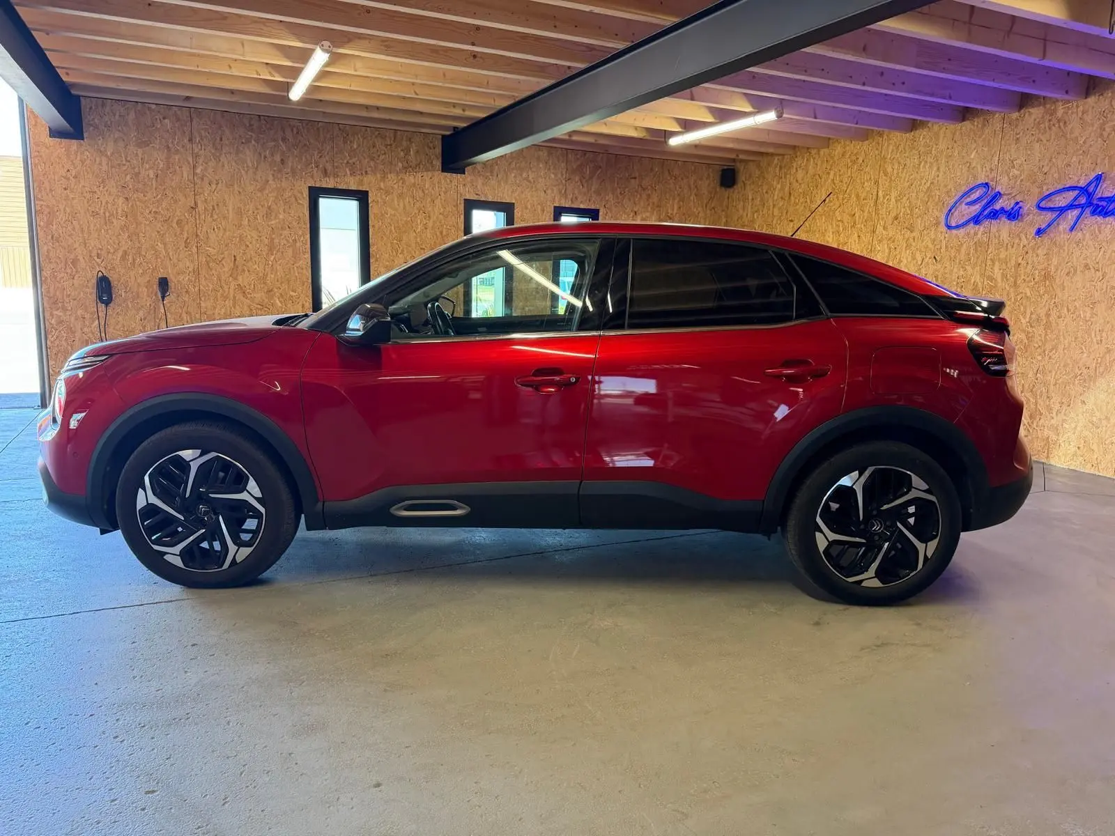 Vue de profil côté gauche d'une Citroën C4 X rouge brillant avec jantes noires et toit noir dans un garage.