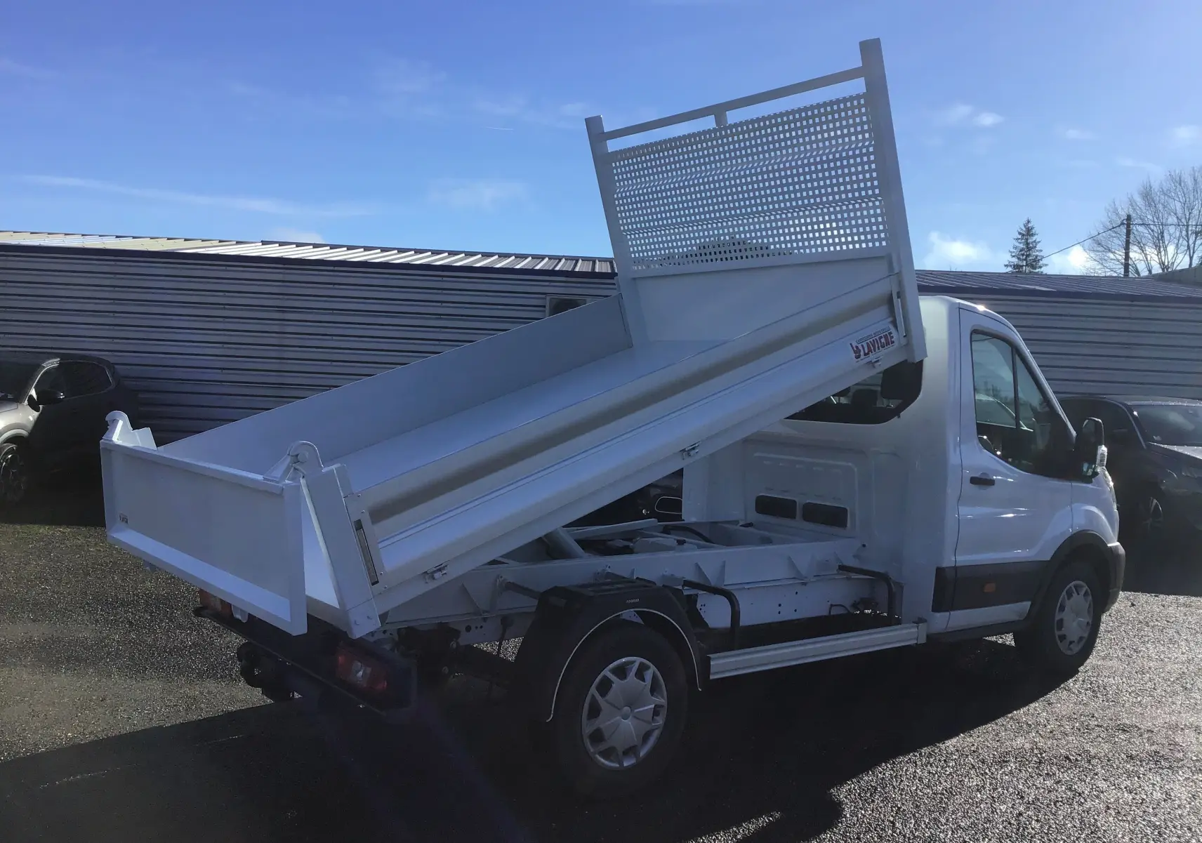 Vue 3/4 arrière droite d'un Ford Transit blanc avec benne basculante relevée sur un parking extérieur.