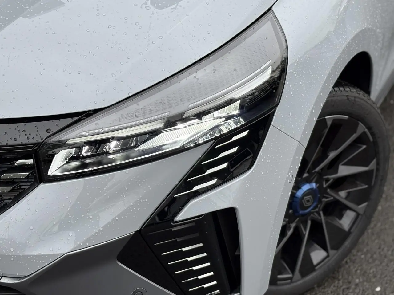 Gros plan sur le phare avant droit et la jante alliage noire avec cabochon bleu d'une Renault Clio Gris Rafale 2025.