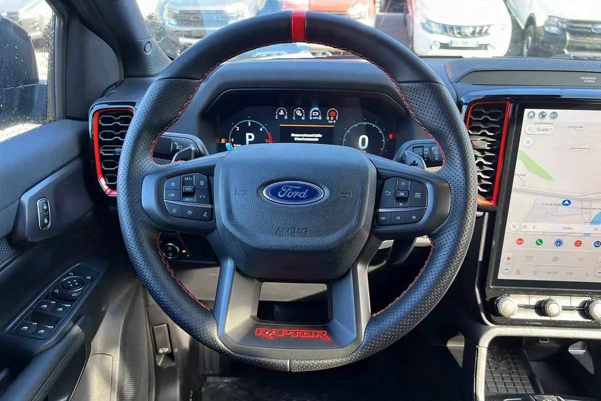 Vue intérieure centrée sur le volant noir perforé avec logo Ford et inscription rouge Raptor, tableau de bord digital et écran tactile.