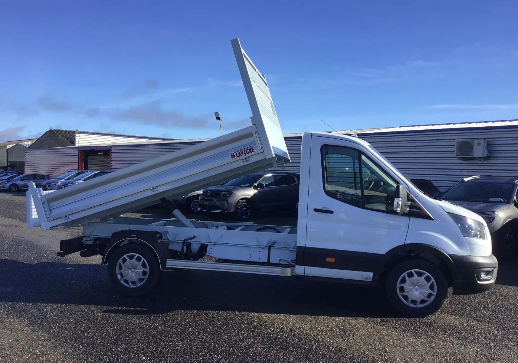 Vue de profil côté droit d'un Ford Transit blanc avec benne basculante levée sur un parking extérieur.