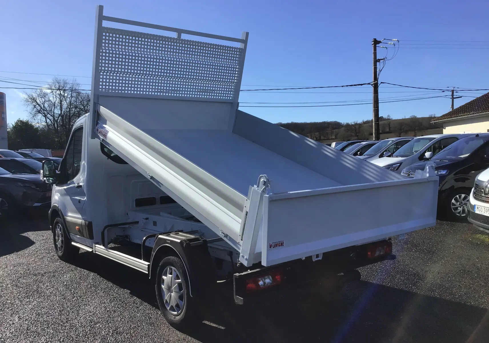 Vue 3/4 arrière droite d'un Ford Transit blanc avec benne relevée et grille de protection arrière sur parking.