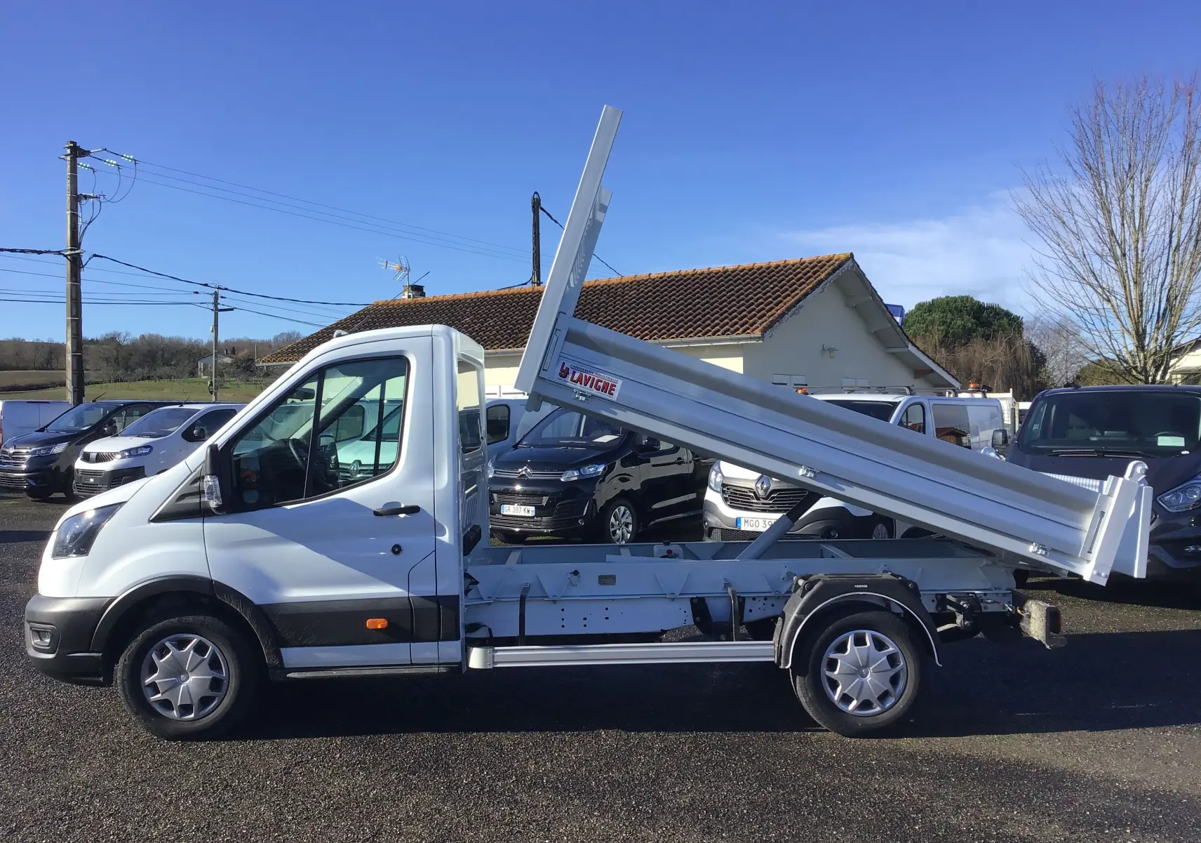 Ford Transit Chassis cabine blanc vu de profil côté gauche avec benne basculante levée sur un parking.