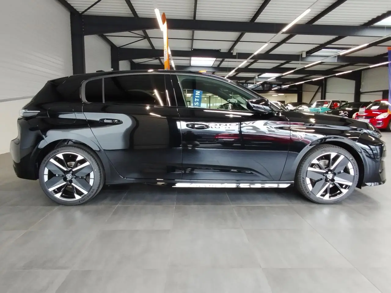 Profil droit d'une Peugeot 308 noire métallisée, avec jantes alliage bi-tons et vitres surteintées en intérieur showroom.