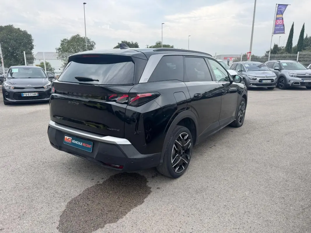 Vue 3/4 arrière droite d’un Peugeot 5008 Hybrid noir avec jantes alliage 19 pouces et barres de toit visibles.
