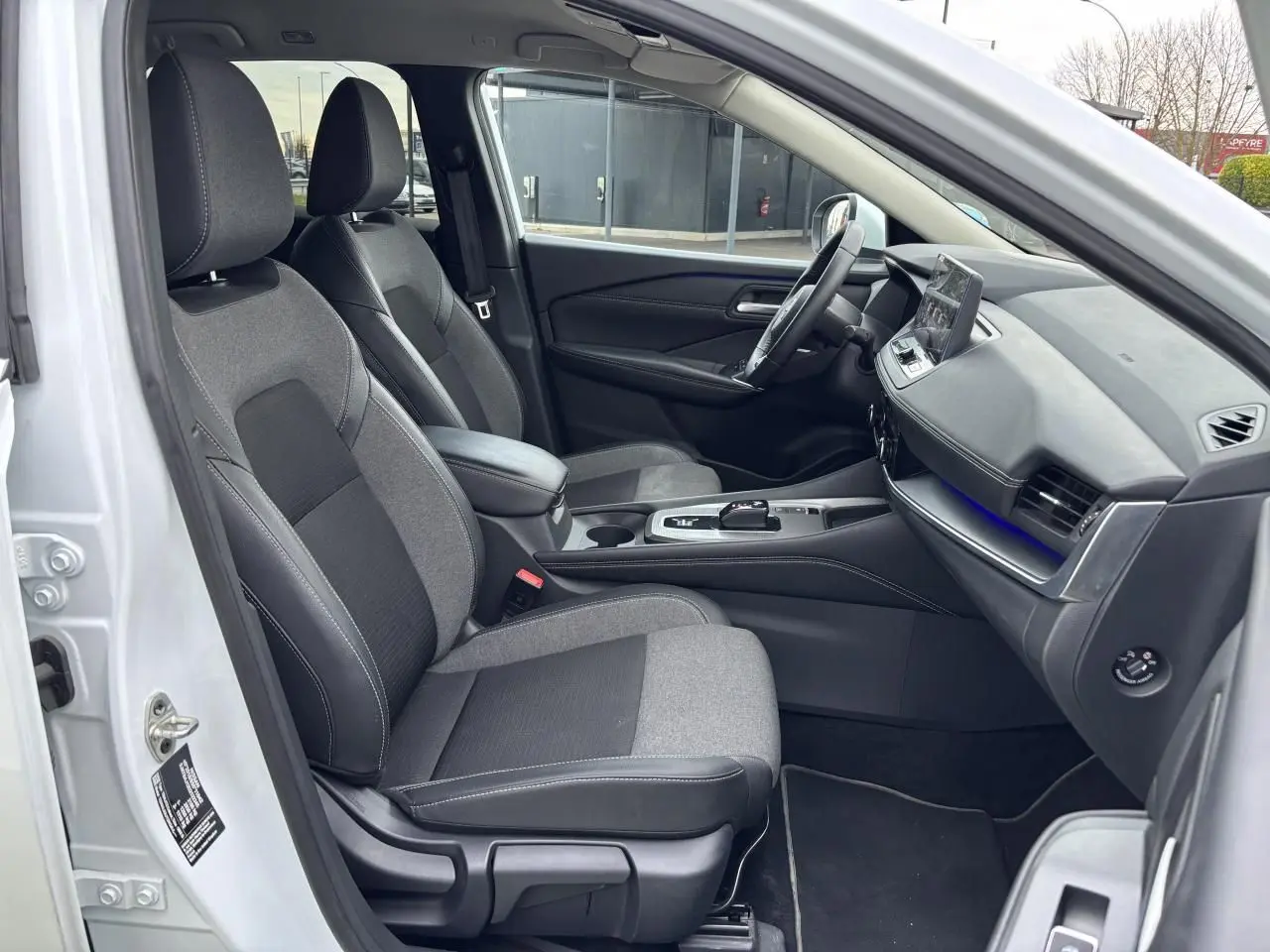 Vue intérieure côté conducteur du Nissan Qashqai blanc nacré 2025, sièges tissu noir et gris avec console centrale moderne.