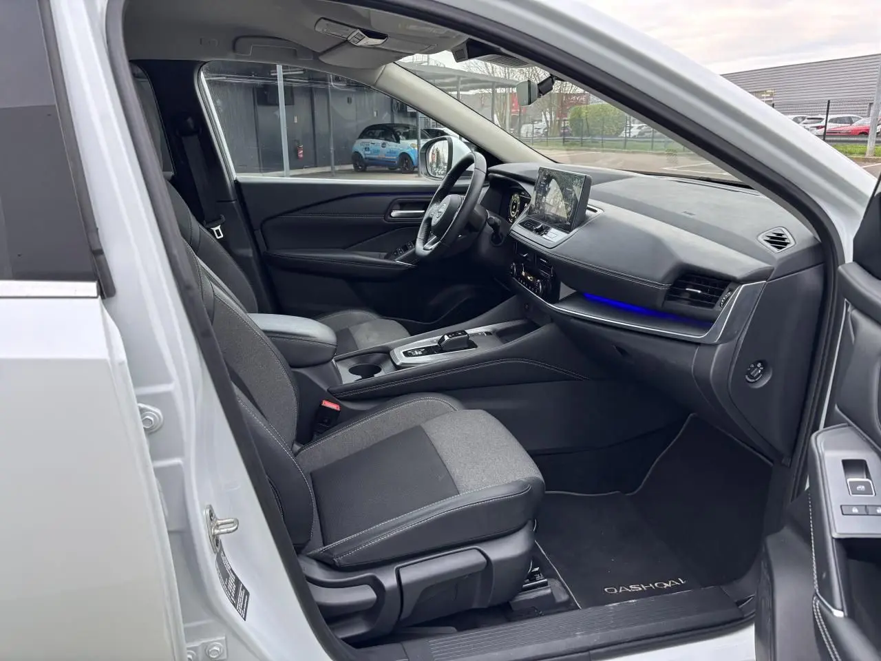 Vue intérieure côté conducteur du Nissan Qashqai blanc nacré 2025, avec tableau de bord moderne et éclairage d'ambiance bleu.