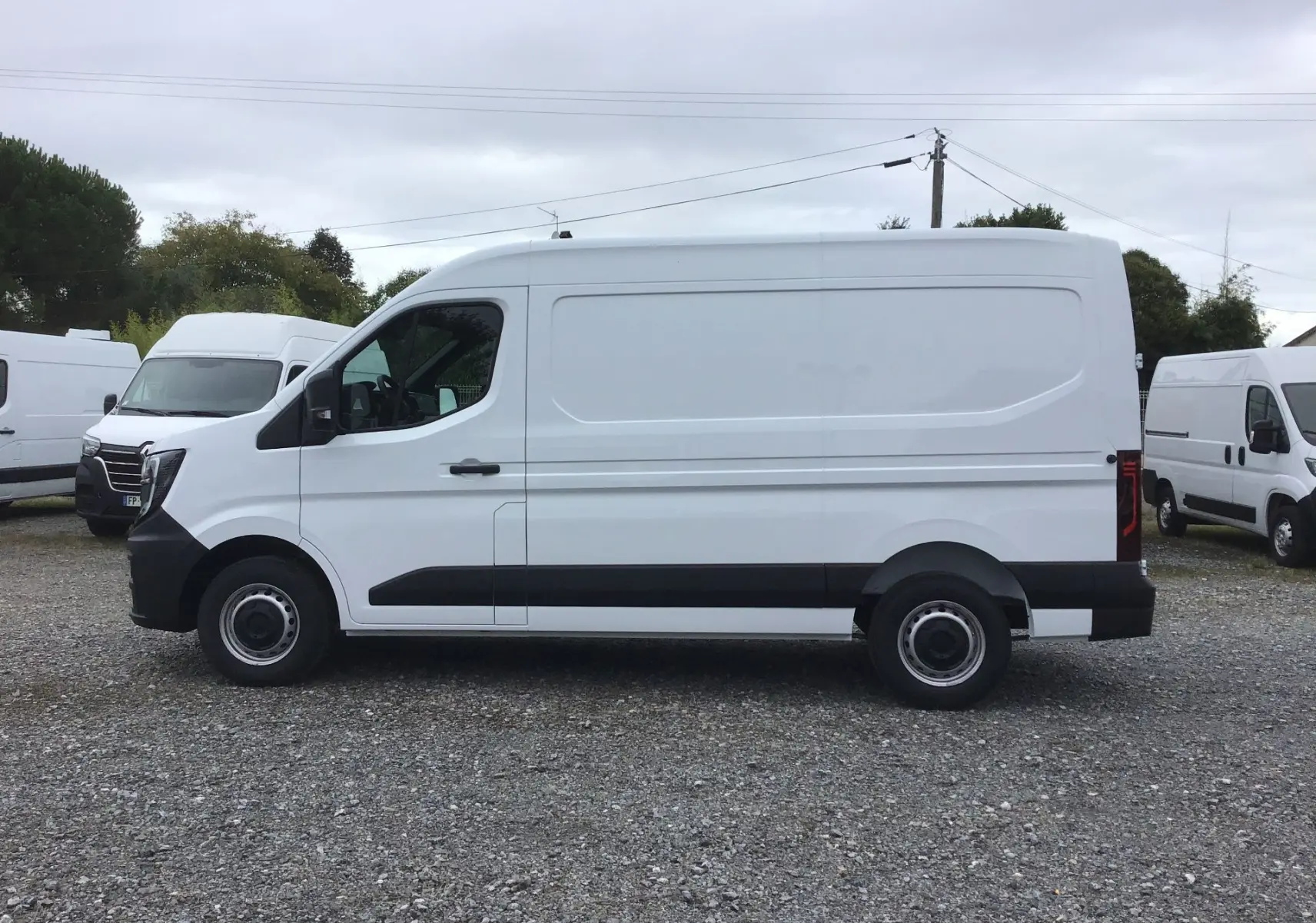 Vue de profil côté gauche d’un utilitaire blanc Renault Master L2H2 avec bandeau noir sur bas de caisse, stationné sur sol gravillonné.
