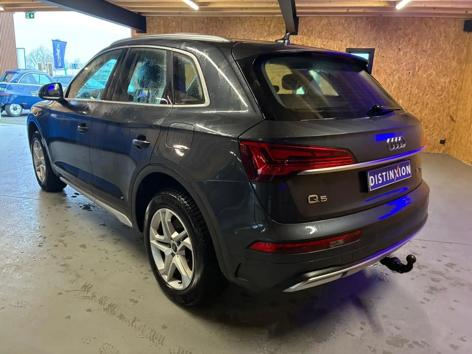 Audi Q5 gris en vue 3/4 arrière droit, avec jantes 5 branches doubles et crochet d'attelage escamotable visible.