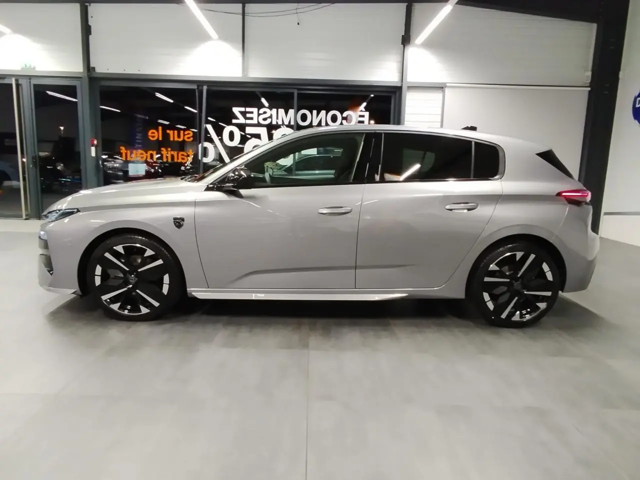 Peugeot 308 Hybrid gris Artense vue de profil côté gauche, jantes alliage noires et toit noir brillant visible en intérieur showroom.