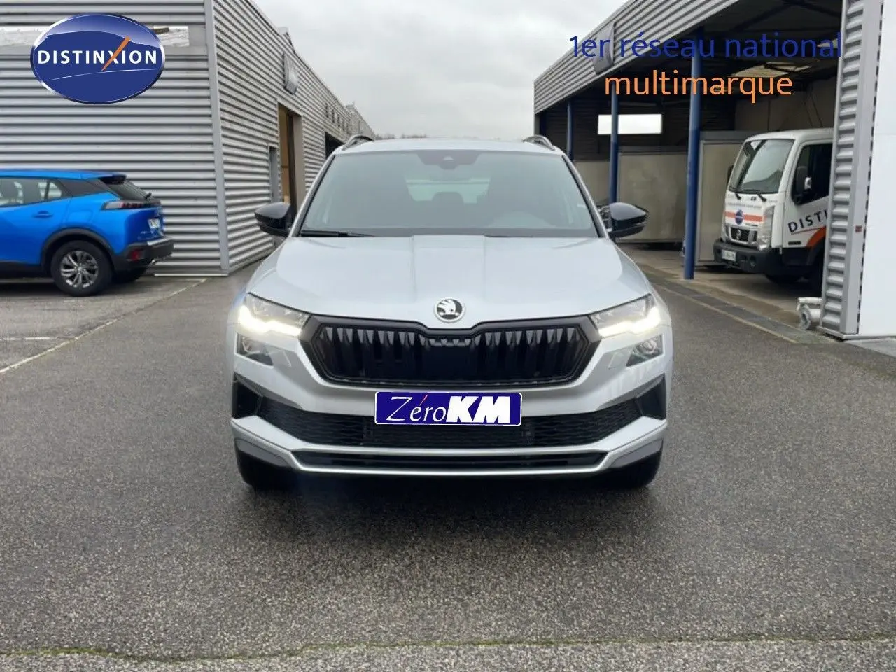Vue frontale d'un Skoda Karoq gris argent métal 2025 avec calandre noire et feux allumés sur parking.