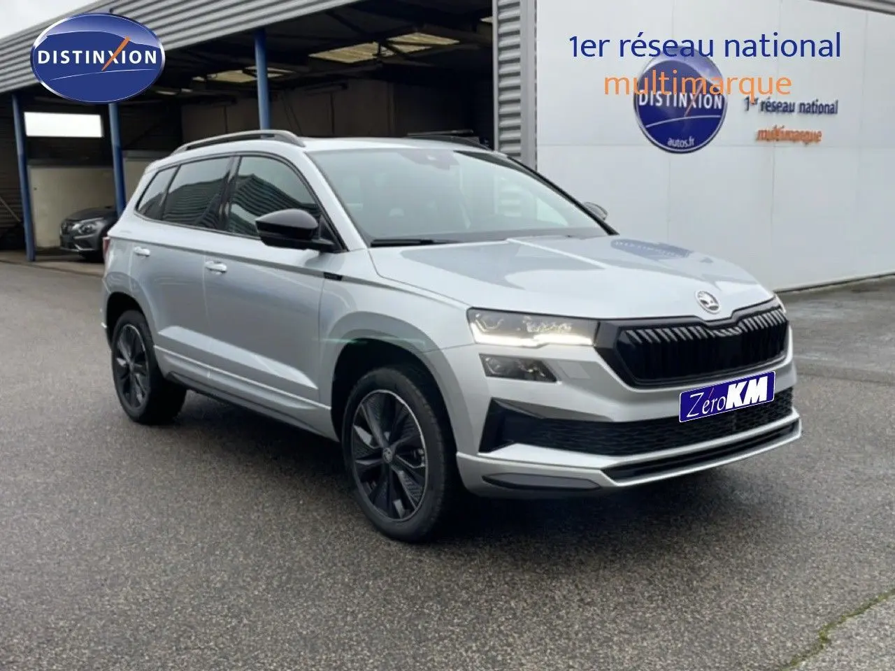 Vue 3/4 avant droite d'un Skoda Karoq gris argent métallisé avec calandre noire et jantes foncées, stationné devant un bâtiment.