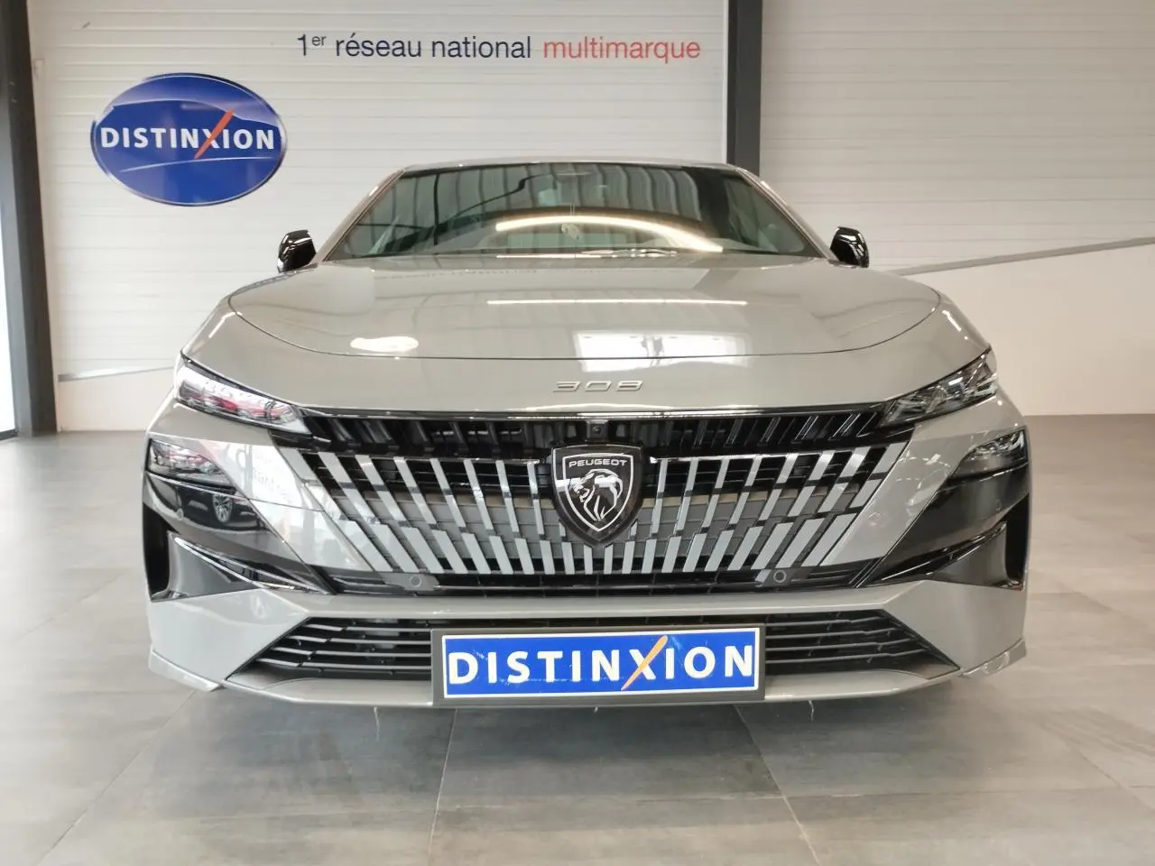 Vue frontale d'une Peugeot 308 gris selenium avec calandre noire et blason Peugeot lumineux, en intérieur showroom.