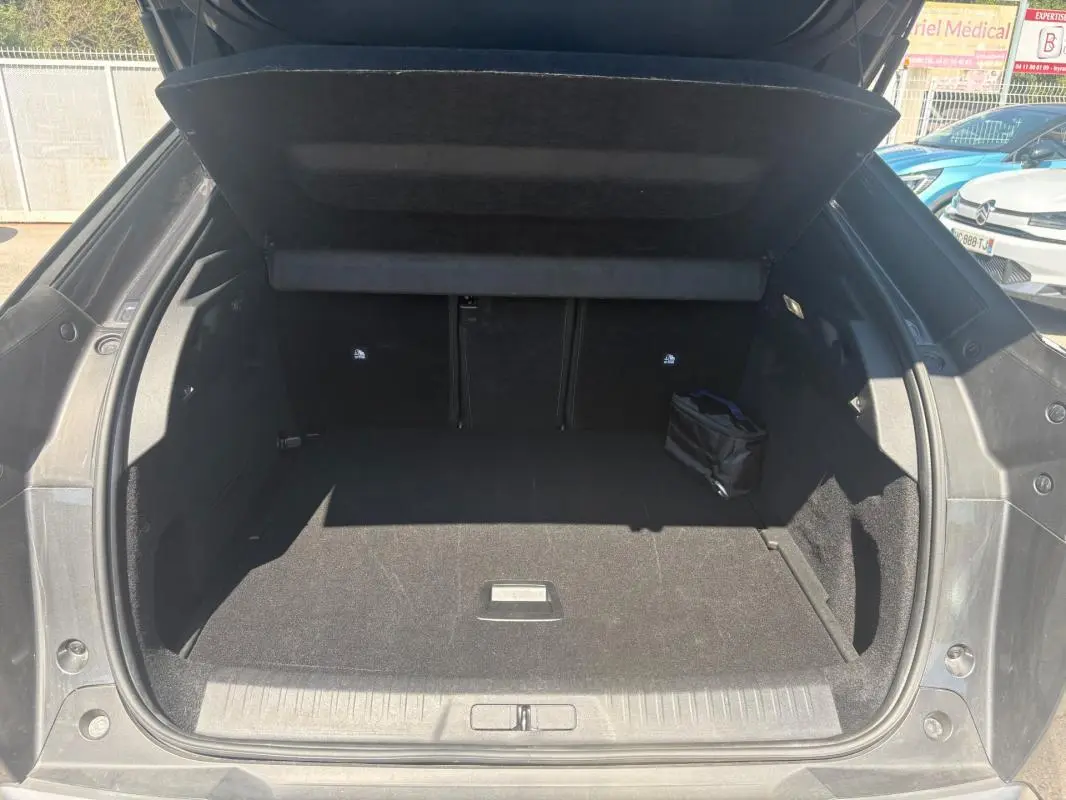 Coffre ouvert vu de l'arrière d'un Peugeot 3008 Hybrid gris, espace de chargement vide avec petit sac noir à droite.
