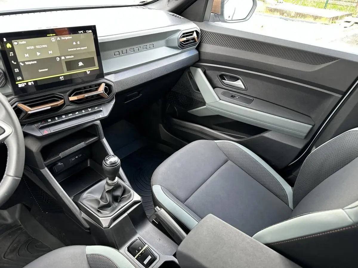 Vue intérieure côté passager du Dacia Duster 2025, tableau de bord noir avec écran tactile et levier de vitesses manuel.