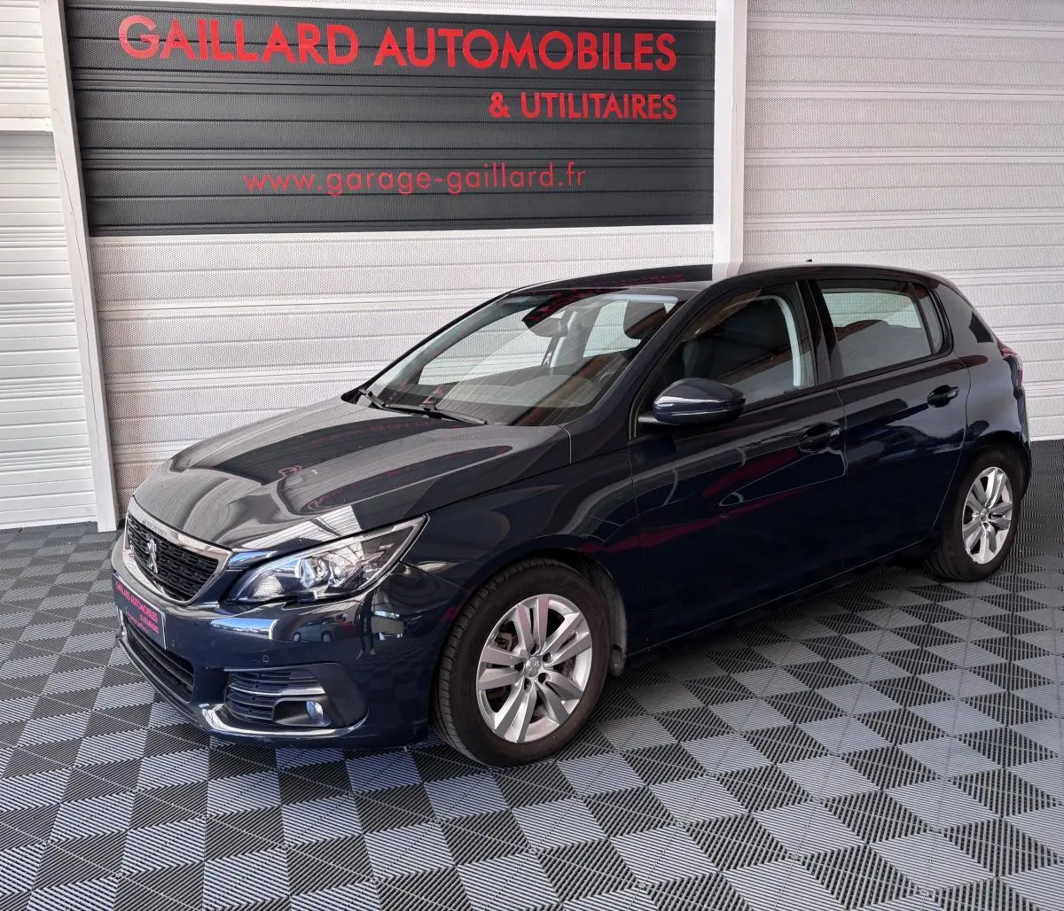 Peugeot 308 gris foncé vue 3/4 avant droit, stationnée en intérieur sur sol damier avec enseigne rouge en arrière-plan.