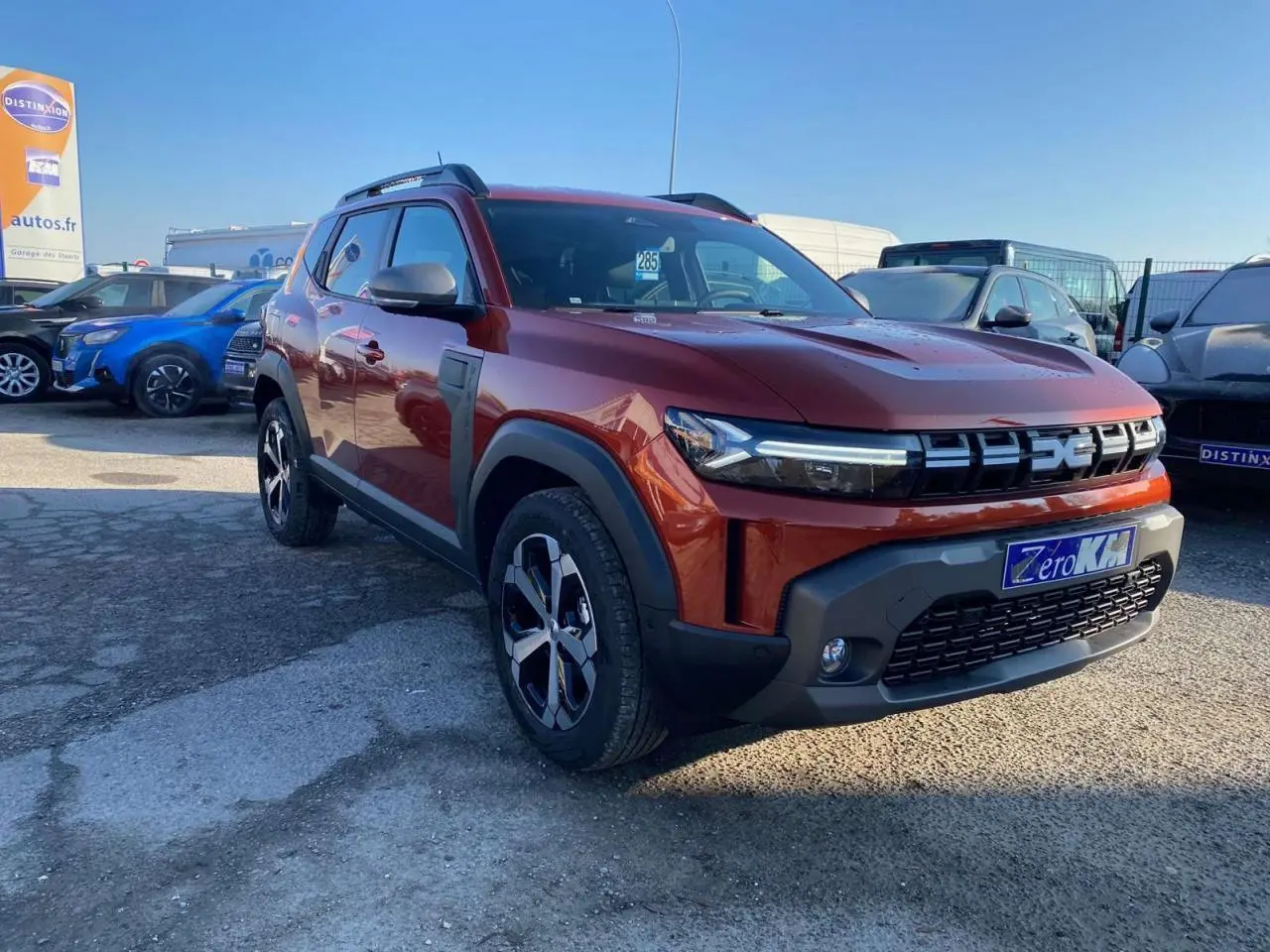 Dacia Duster brun terracotta en 3/4 avant droit, avec jantes alliage 18 pouces et barres de toit noires.