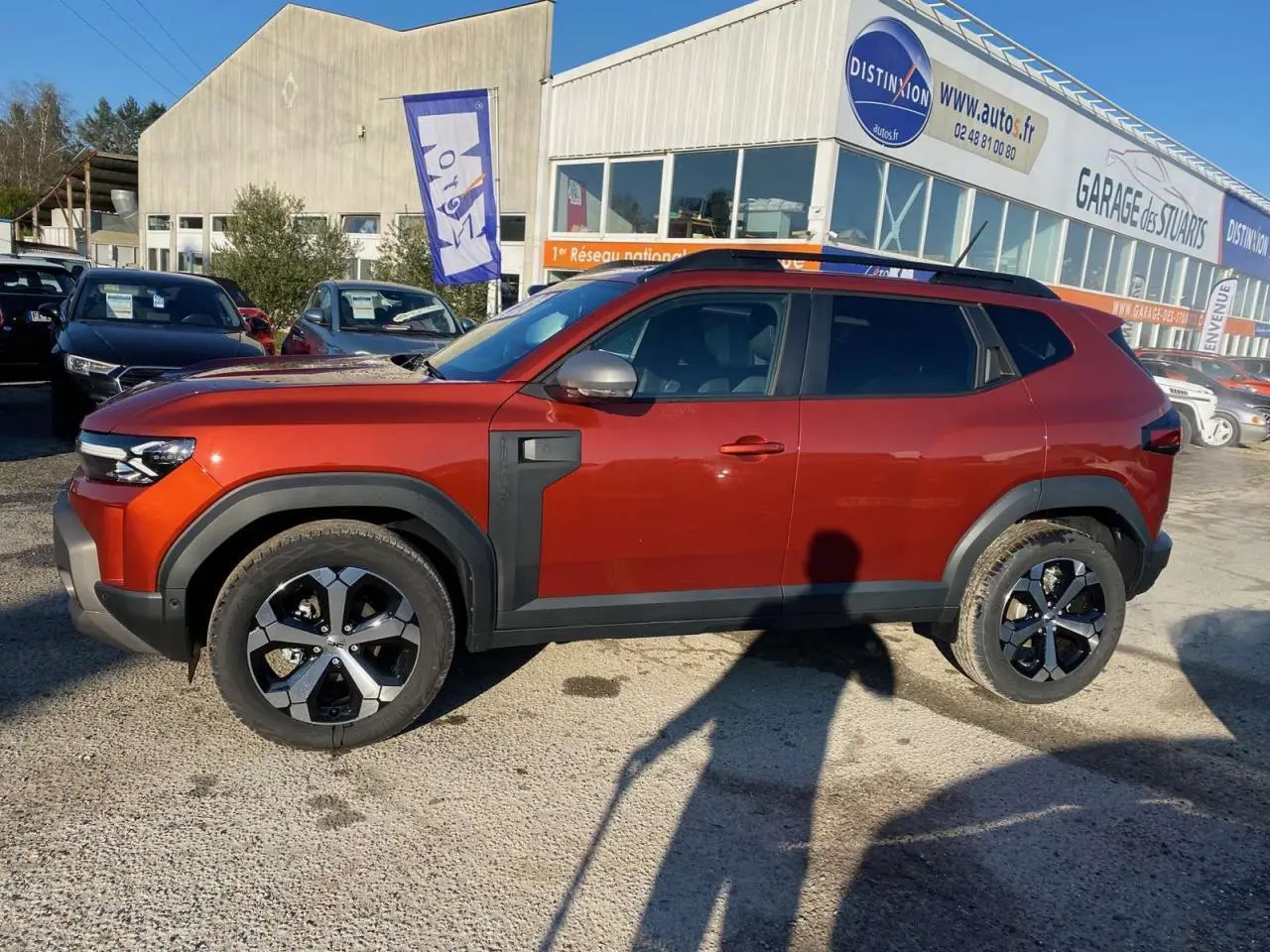 Profil côté gauche du Dacia Duster 2024 brun terracotta avec barres de toit noires et jantes alliage 18 pouces.