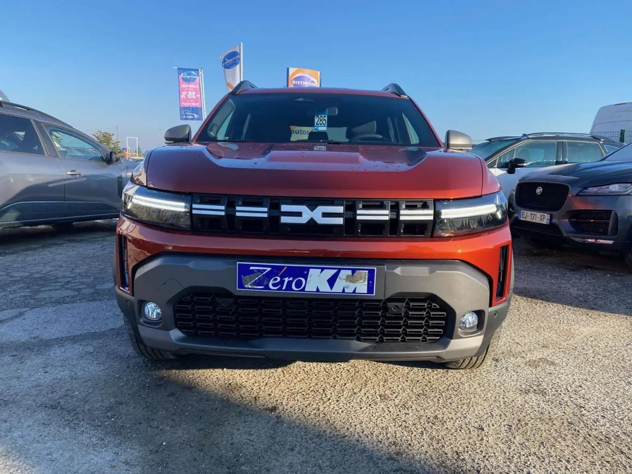 Vue de face d'un Dacia Duster 2024 brun terracotta avec calandre marquée et plaques "Zéro KM" sur un parking extérieur.