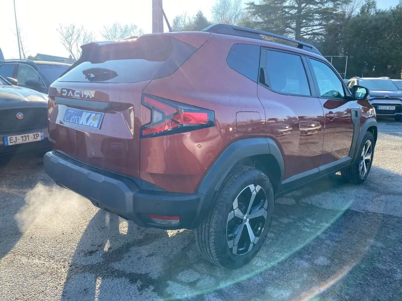 Vue 3/4 arrière droite du Dacia Duster 2024 brun terracotta avec jantes alliage et barres de toit noires.