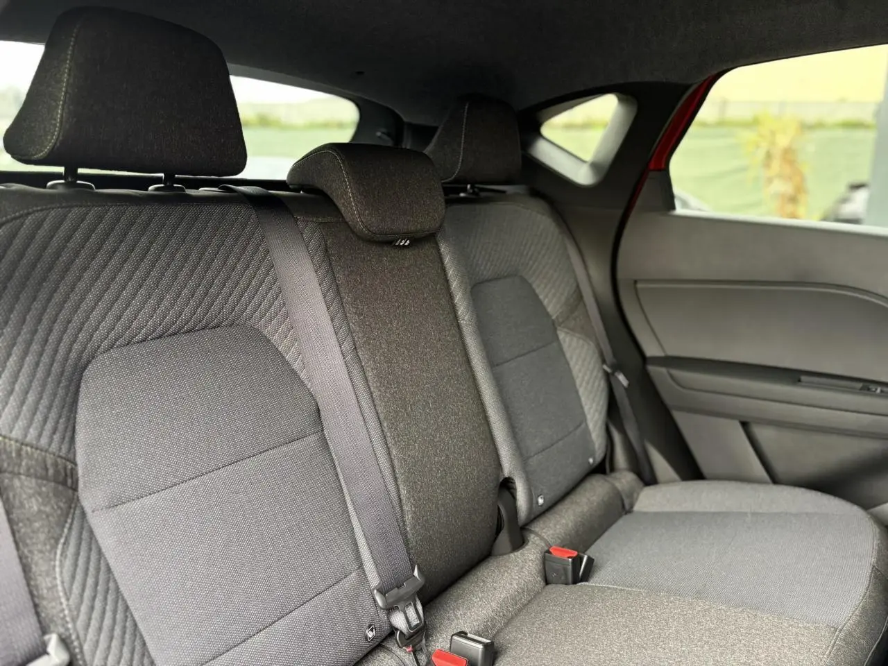 Vue intérieure arrière du Renault Captur 2025, sièges tissu gris avec ceinture de sécurité centrale visible et partie de la porte côté droit rouge Flamme.