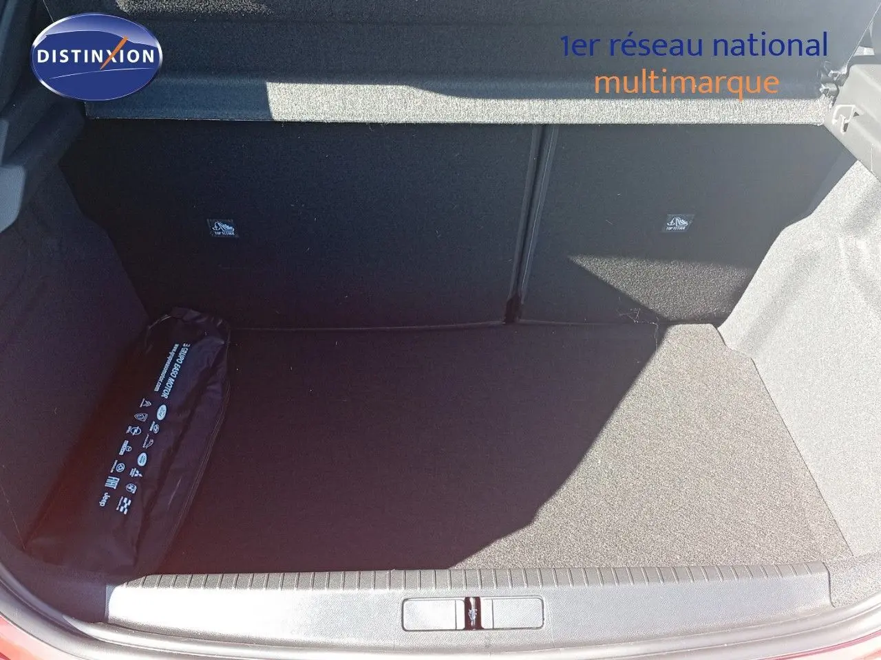 Coffre ouvert d'une Opel Corsa rouge métal avec toit noir, montrant un espace de rangement vide et un kit de sécurité noir.