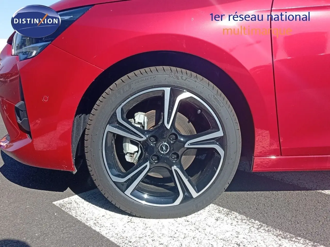 Gros plan sur la roue avant gauche d'une Opel Corsa rouge métal avec jante noire et détails argentés.