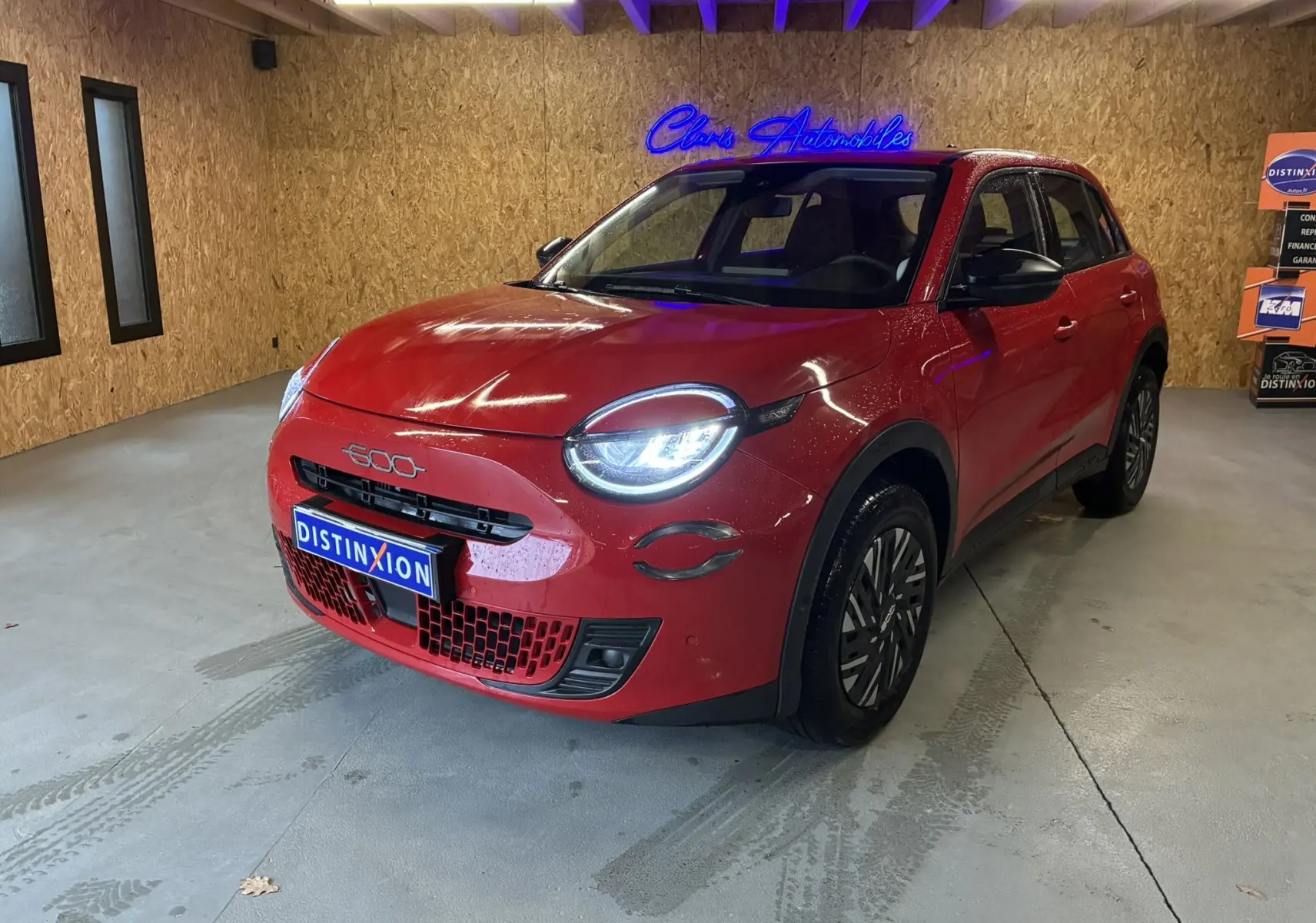 Vue 3/4 avant d'une FIAT 600 rouge 2024 avec phares allumés, calandre noire et jantes stylisées en intérieur bois.