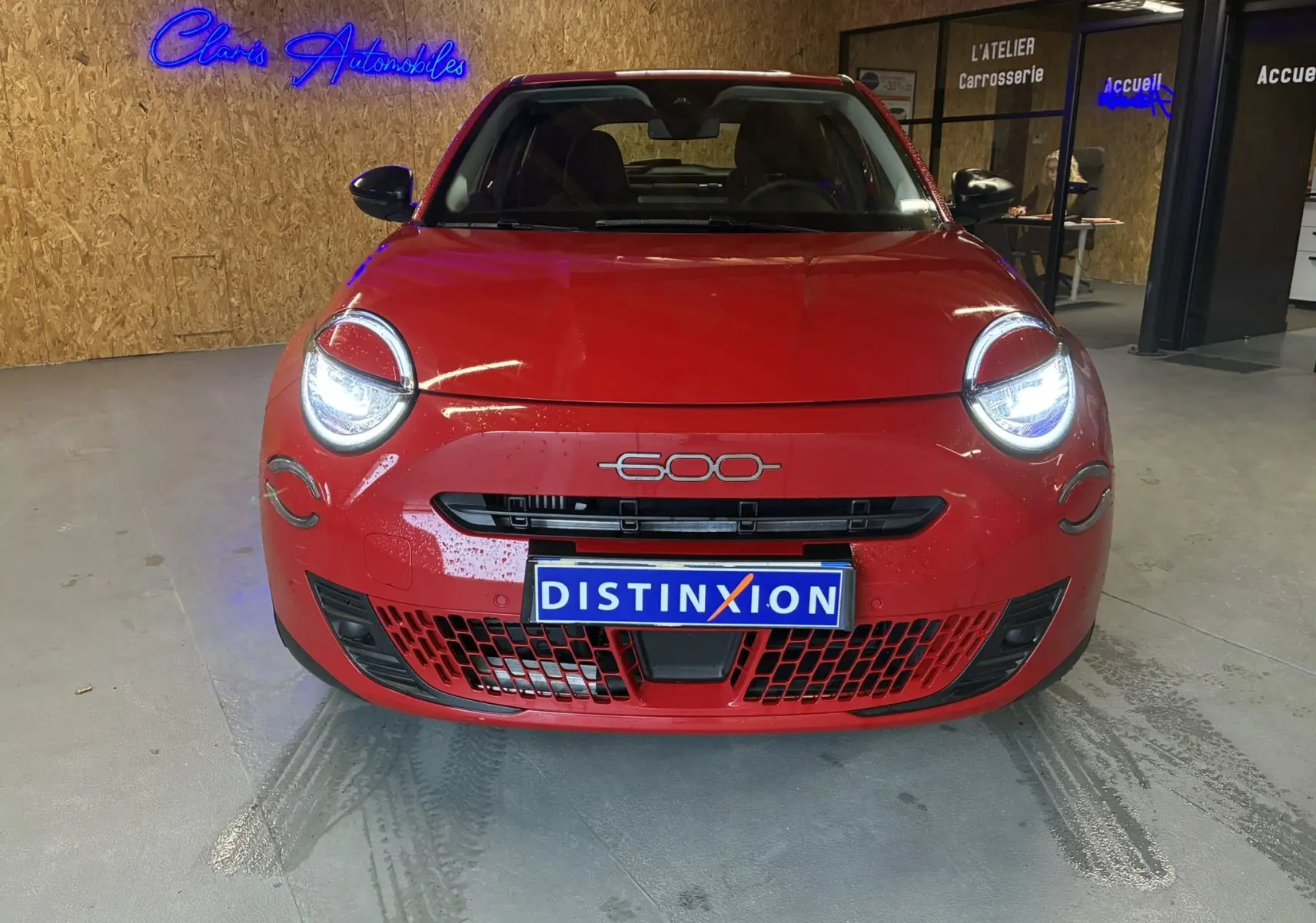 Vue frontale d'une FIAT 600 rouge 2024 avec phares allumés, plaque Distinxion et ambiance intérieure d'un showroom.