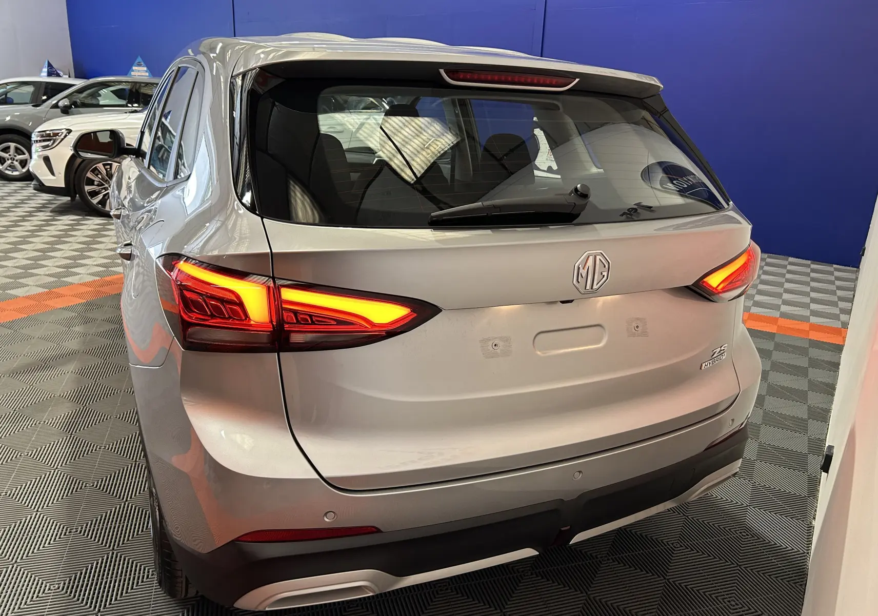 Vue arrière en 3/4 droit du MG ZS Cosmic Silver avec feux allumés dans un showroom intérieur.