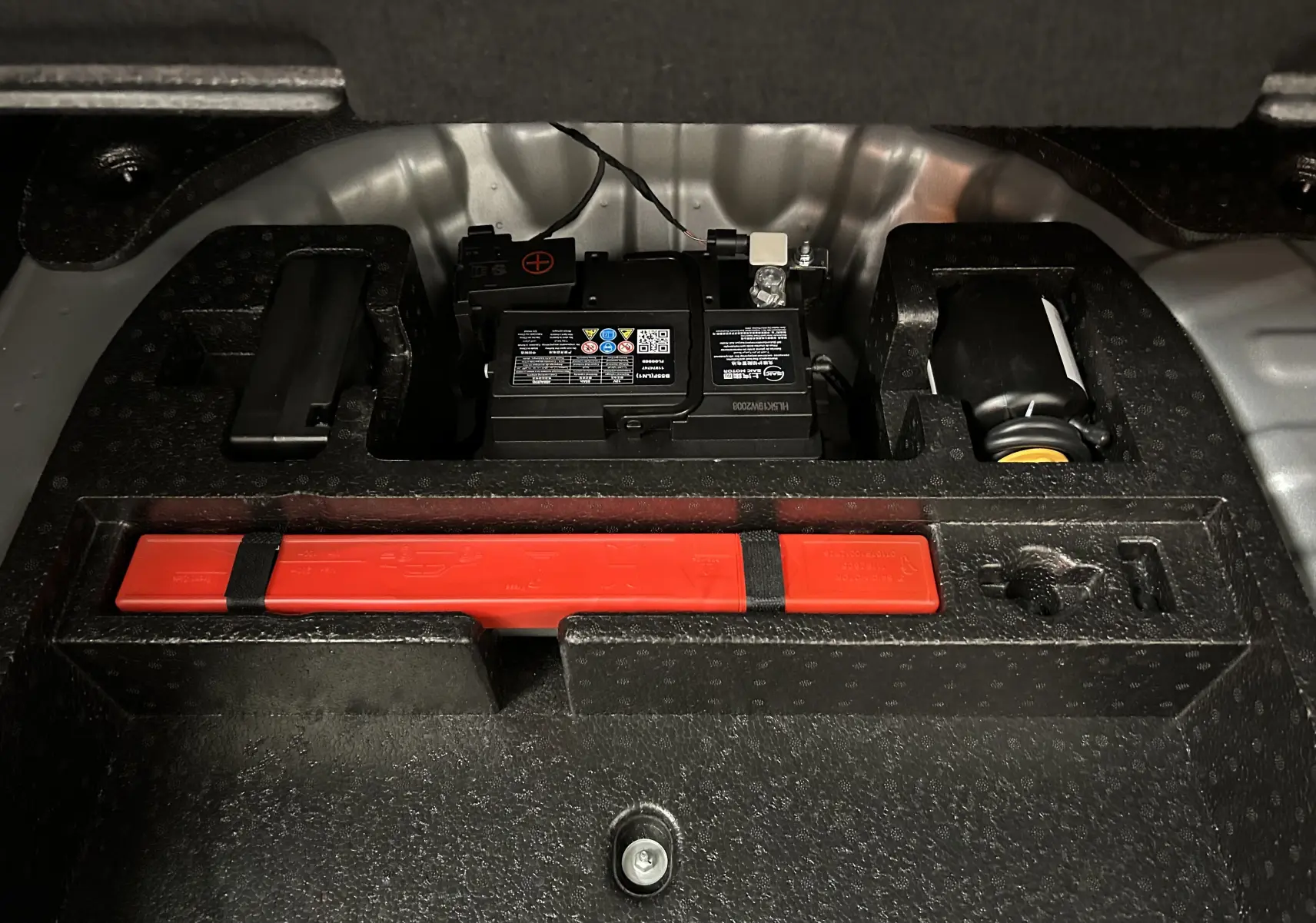 Vue rapprochée du compartiment de rangement sous le coffre du MG ZS Cosmic Silver, montrant la batterie et un triangle de signalisation rouge.