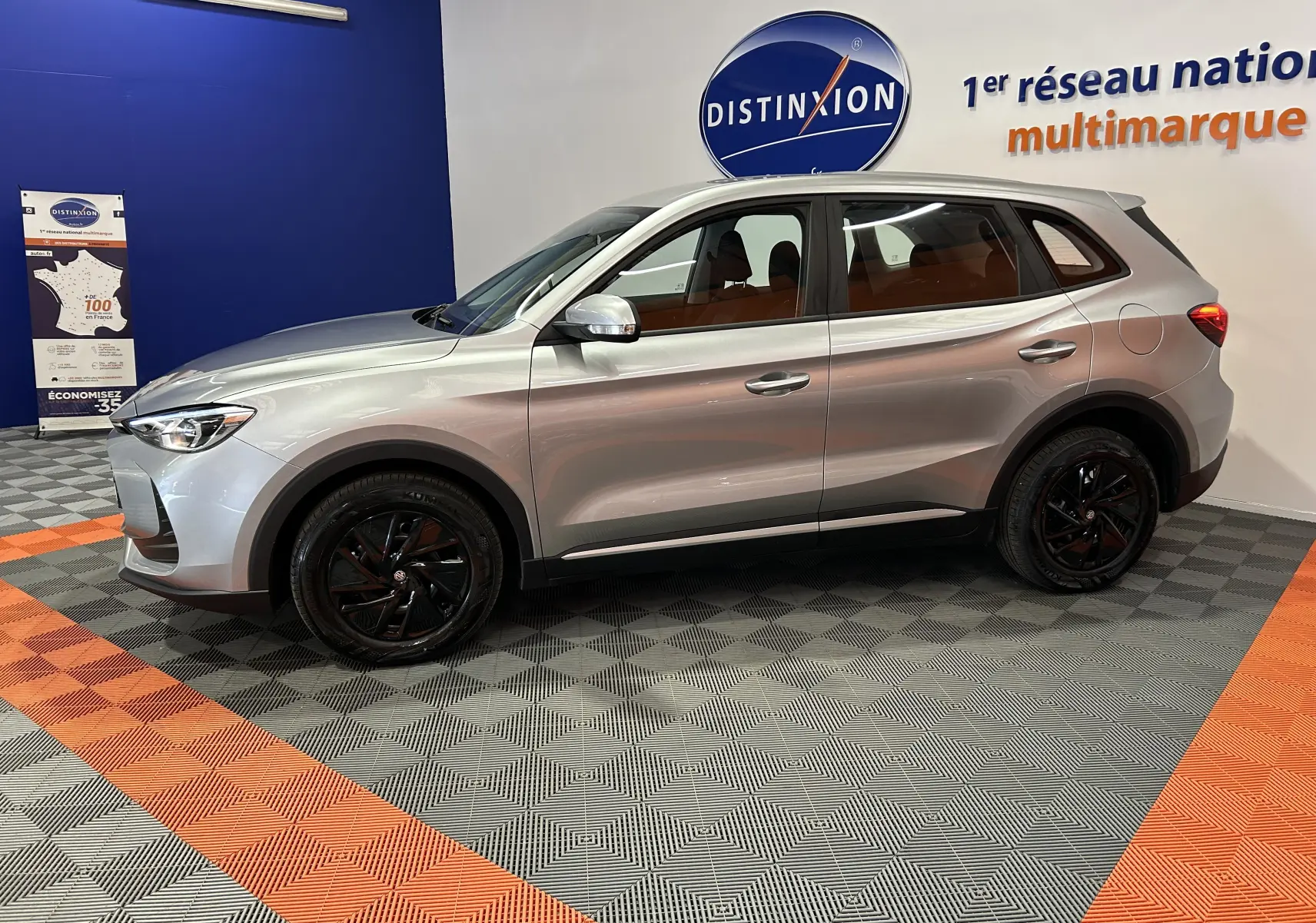 Vue de profil côté gauche du MG ZS 1.5 L Hybrid+ 2025 en Cosmic Silver avec jantes noires, dans un showroom.