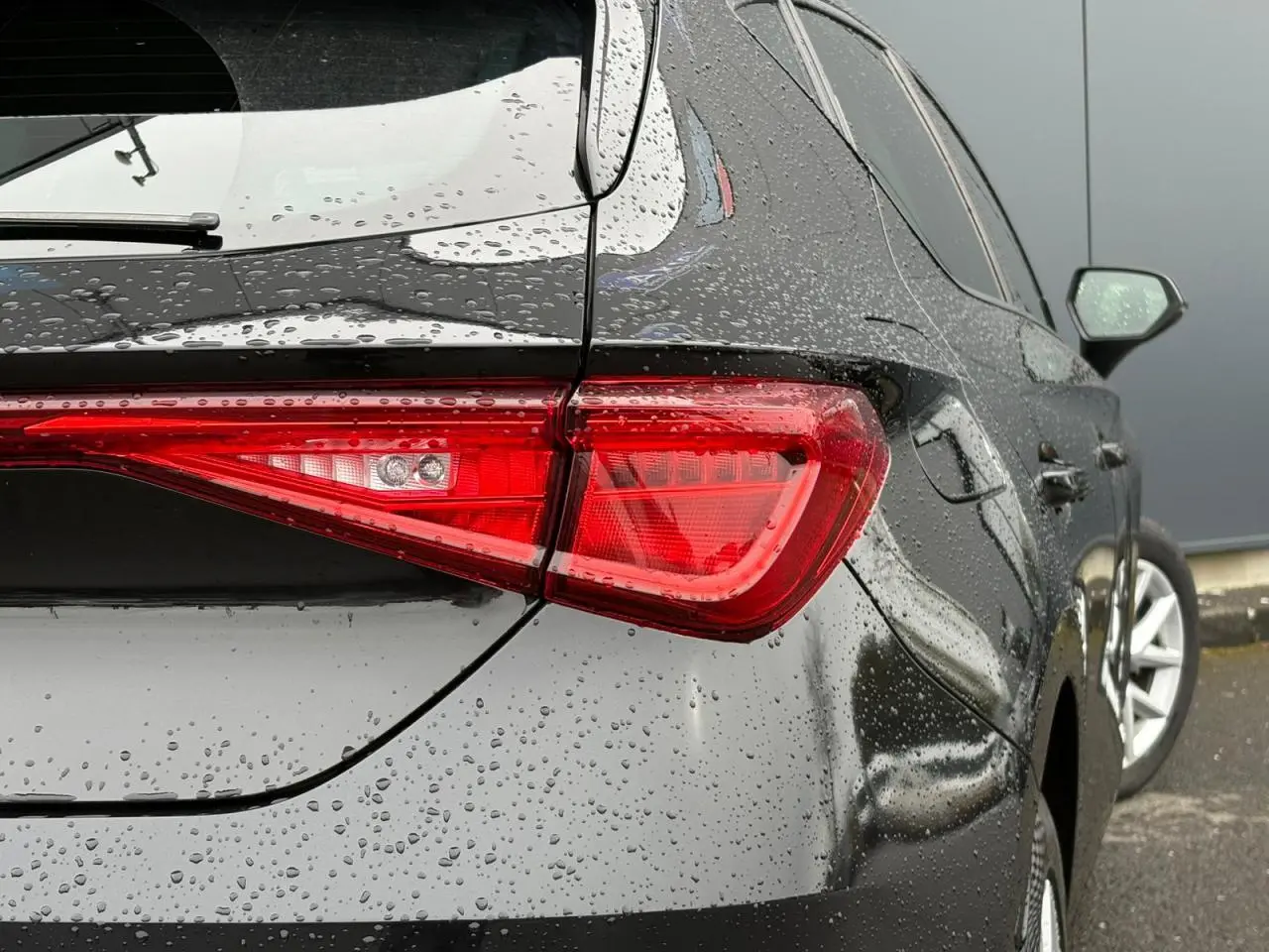 Vue arrière droite d'une SEAT Leon 2024 noir minuit, avec gouttes de pluie sur la carrosserie et feux arrière allumés.