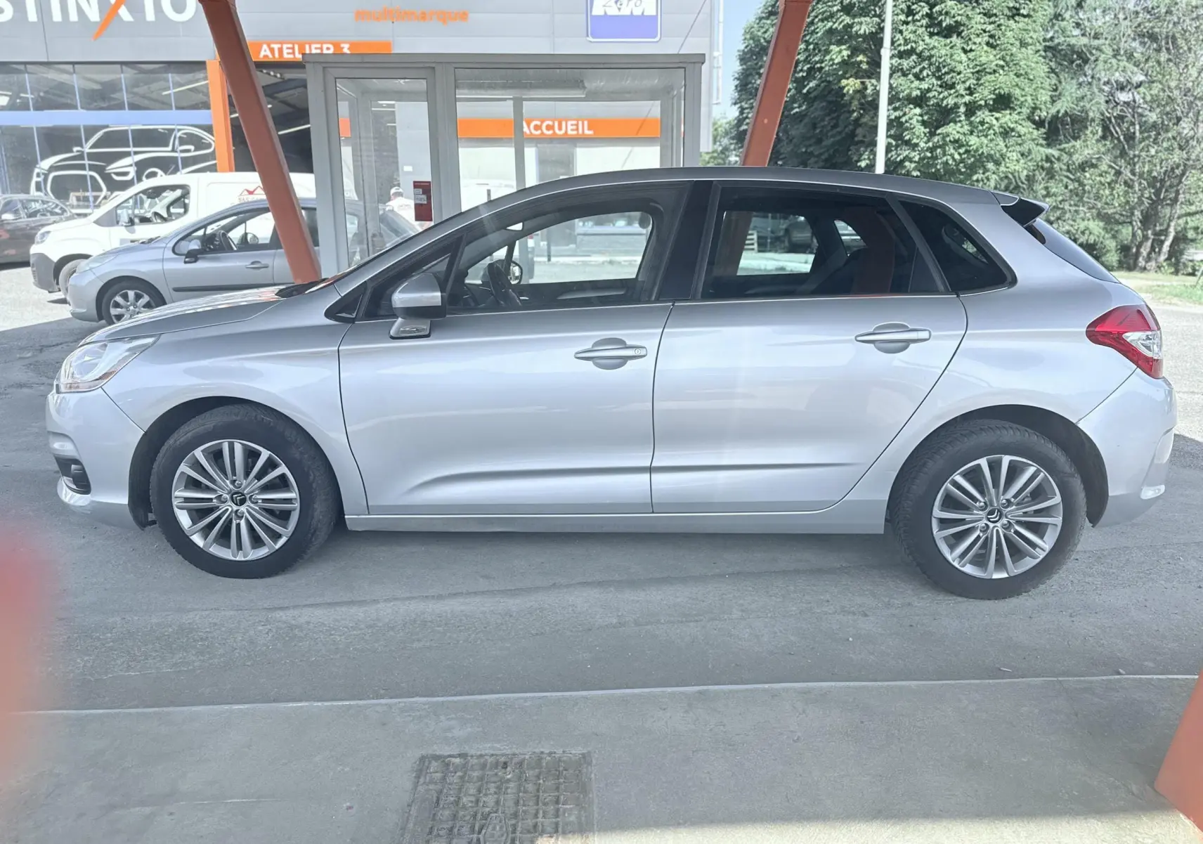 Vue de profil côté gauche d'une Citroën C4 gris métallisé, 5 portes, garée sous un abri avec fond urbain.