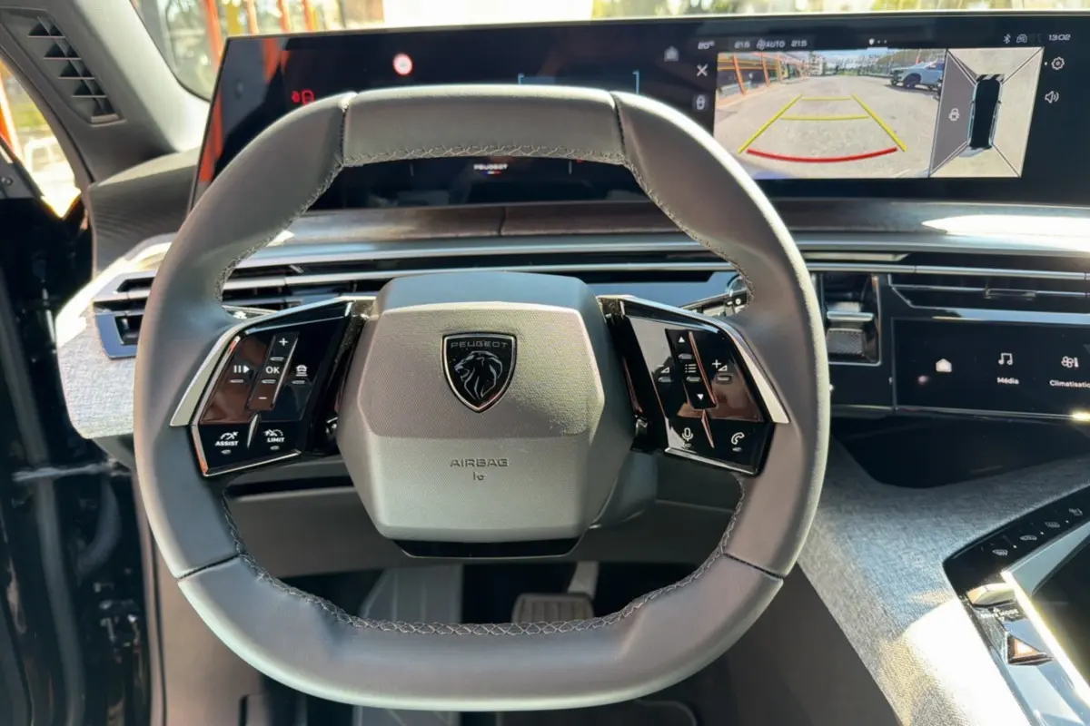 Vue rapprochée du volant cuir multifonctions gris du Peugeot 5008 Hybrid 145 avec écran panoramique affichant la caméra 360° en arrière-plan.