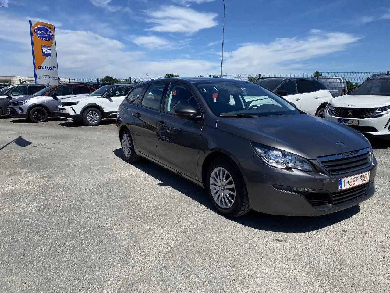 Peugeot 308 SW gris clair vue 3/4 avant droit sur parking ensoleillé avec attelage visible à l'arrière.